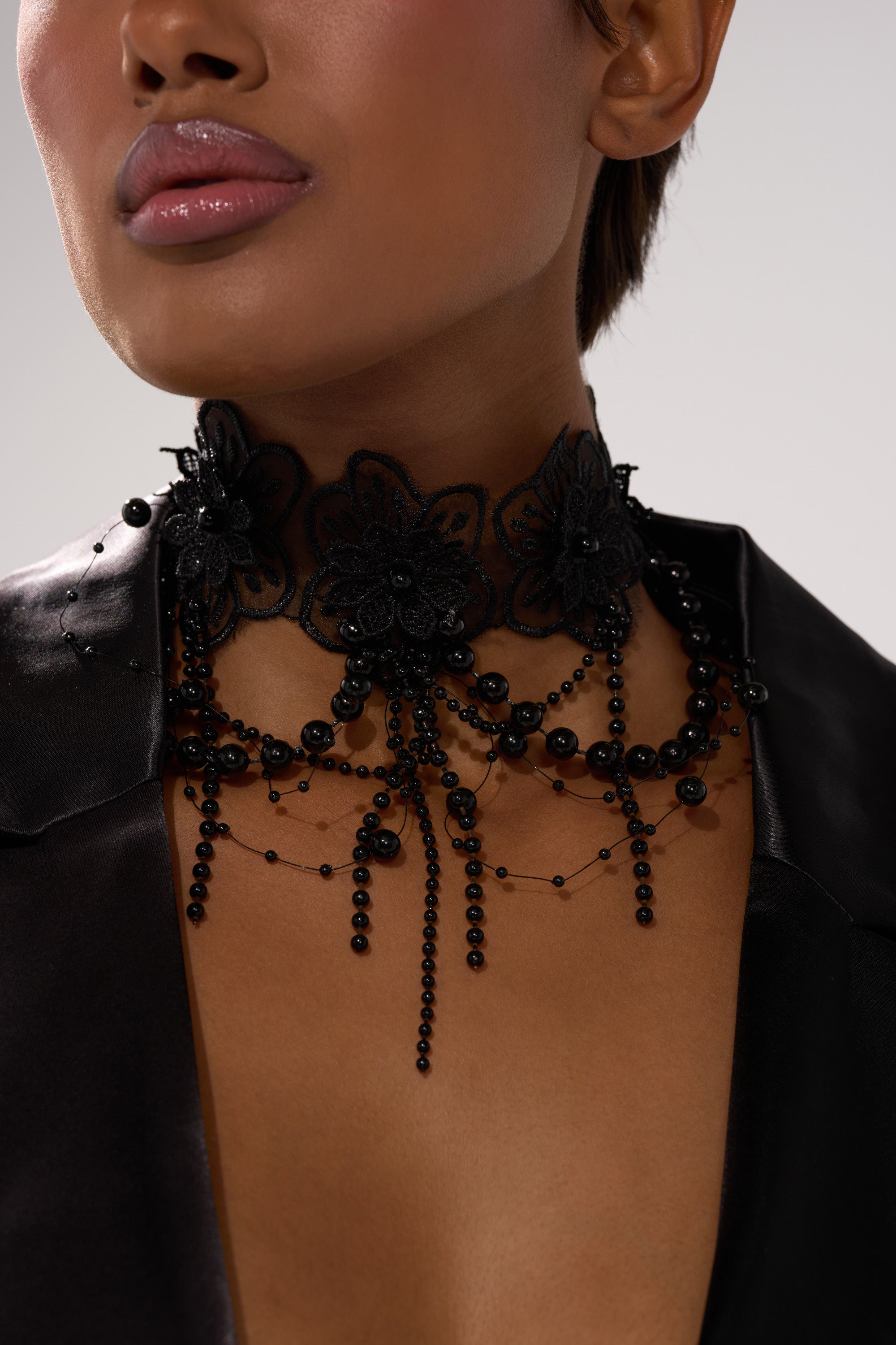 A close-up of a person wearing the LIFE OF THE PARTY NECKLACE, featuring black lace with floral designs and black beaded strands, paired with a black satin garment. The person's lips are glossy against a neutral background. 1