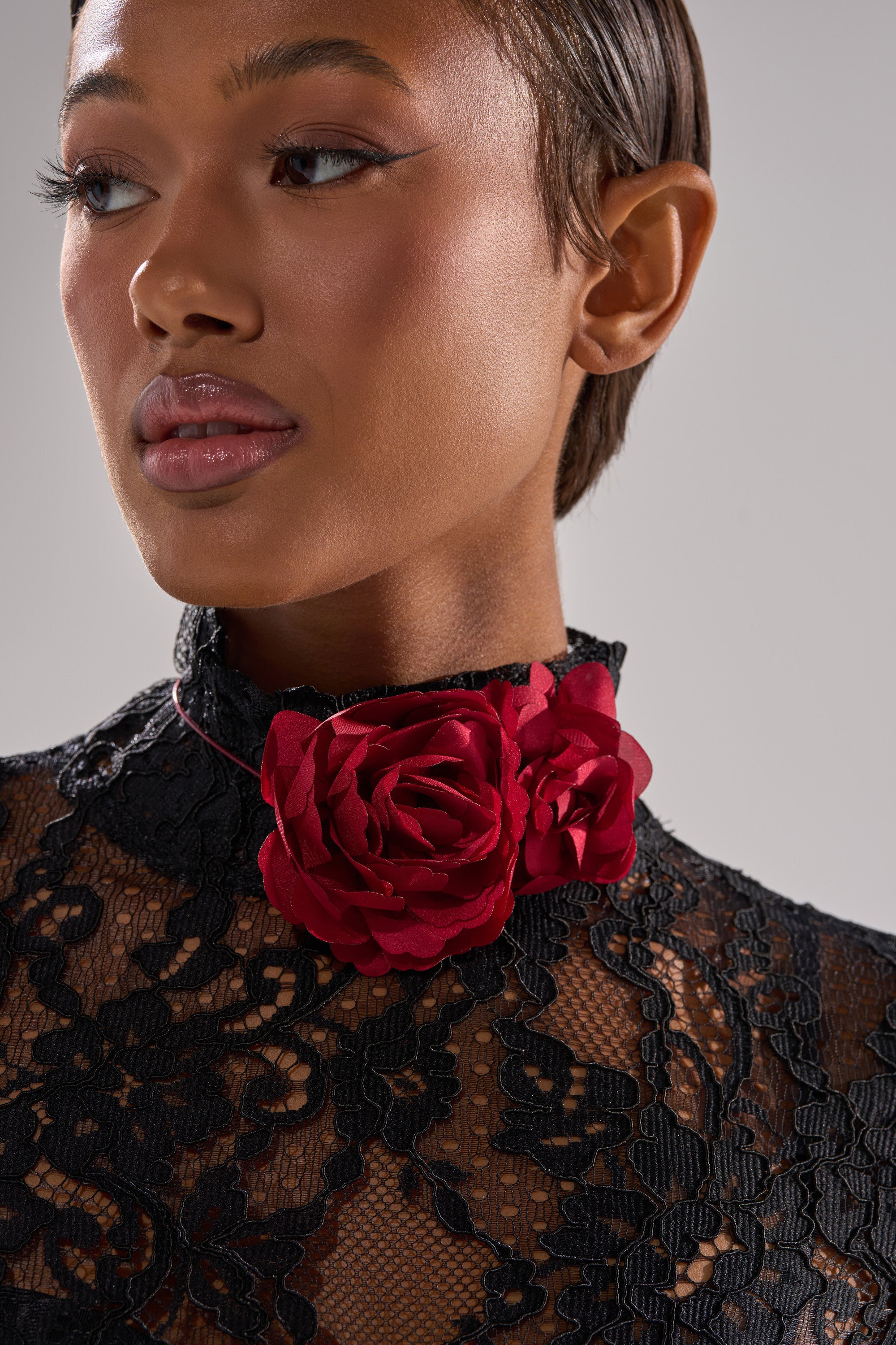 A woman models the ROSEY FLORAL CHOKER NECKLACE, featuring two red fabric roses, paired with a black lace top. She is shown from the shoulders up, gazing to the side against a plain background. 1