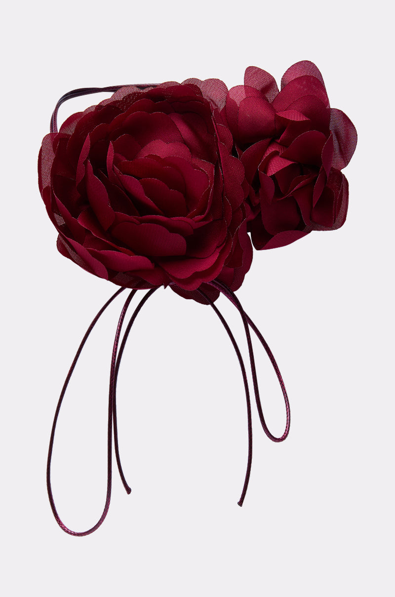 The ROSEY FLORAL CHOKER NECKLACE is styled with a headband featuring two large deep red fabric flowers and long matching ribbons, creating a romantic look against a light gray background. 2