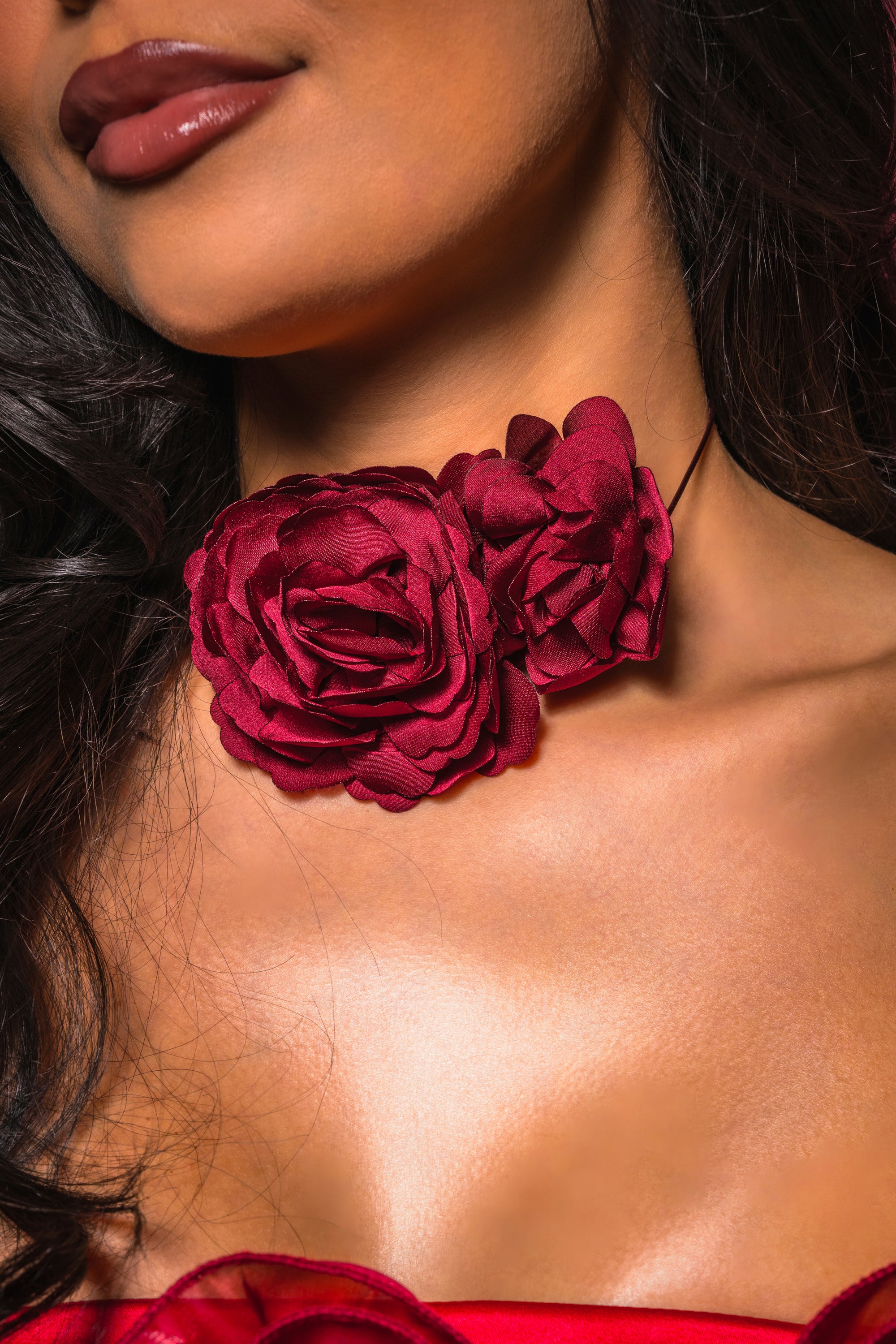 A woman wears the ROSEY FLORAL CHOKER NECKLACE, featuring two lifelike red fabric roses. Only her lower face, neck, and chest are shown, highlighting this bold accessory—perfect for a standout date-night look. 0