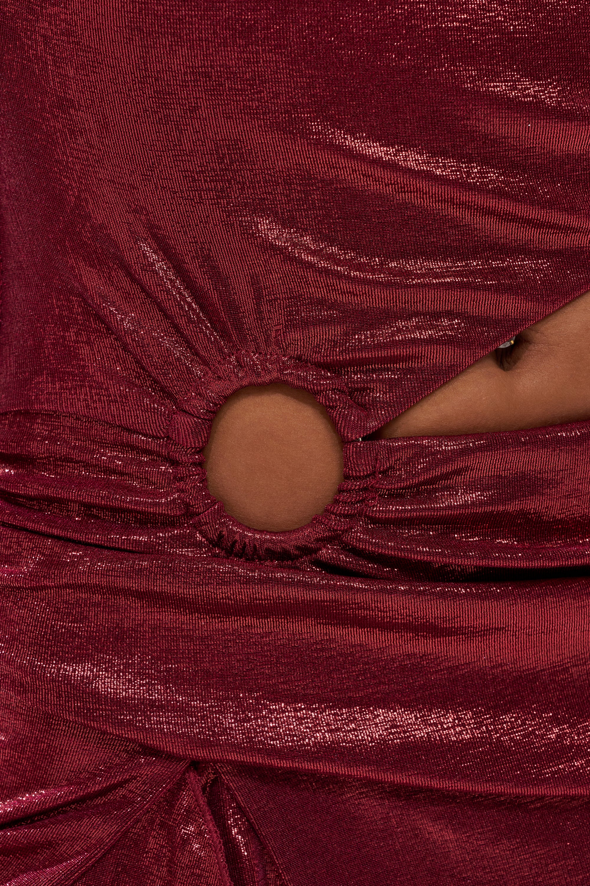 A close-up of the GIRL ON FIRE MINI DRESS—this shiny burgundy metallic mini features a circular waist cutout revealing a hint of skin and part of the belly button. 8