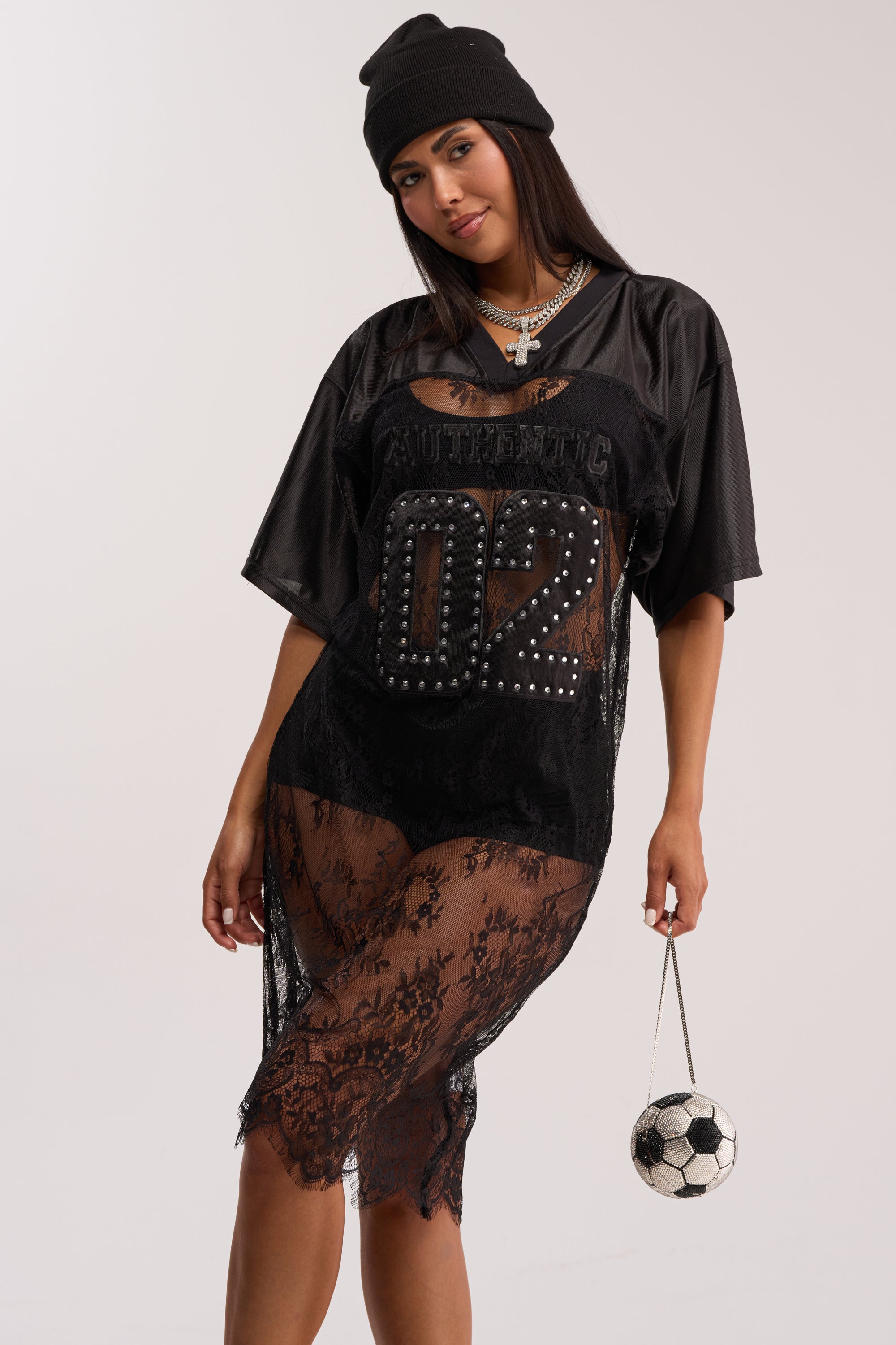 Woman in the PART TIME PLAYER LACE JERSEY MIDI DRESS IN BLACK with "02" and "AUTHENTIC" lettering, paired with a black beanie, layered necklaces, and a soccer ball purse—an edgy festival look against a plain light background.