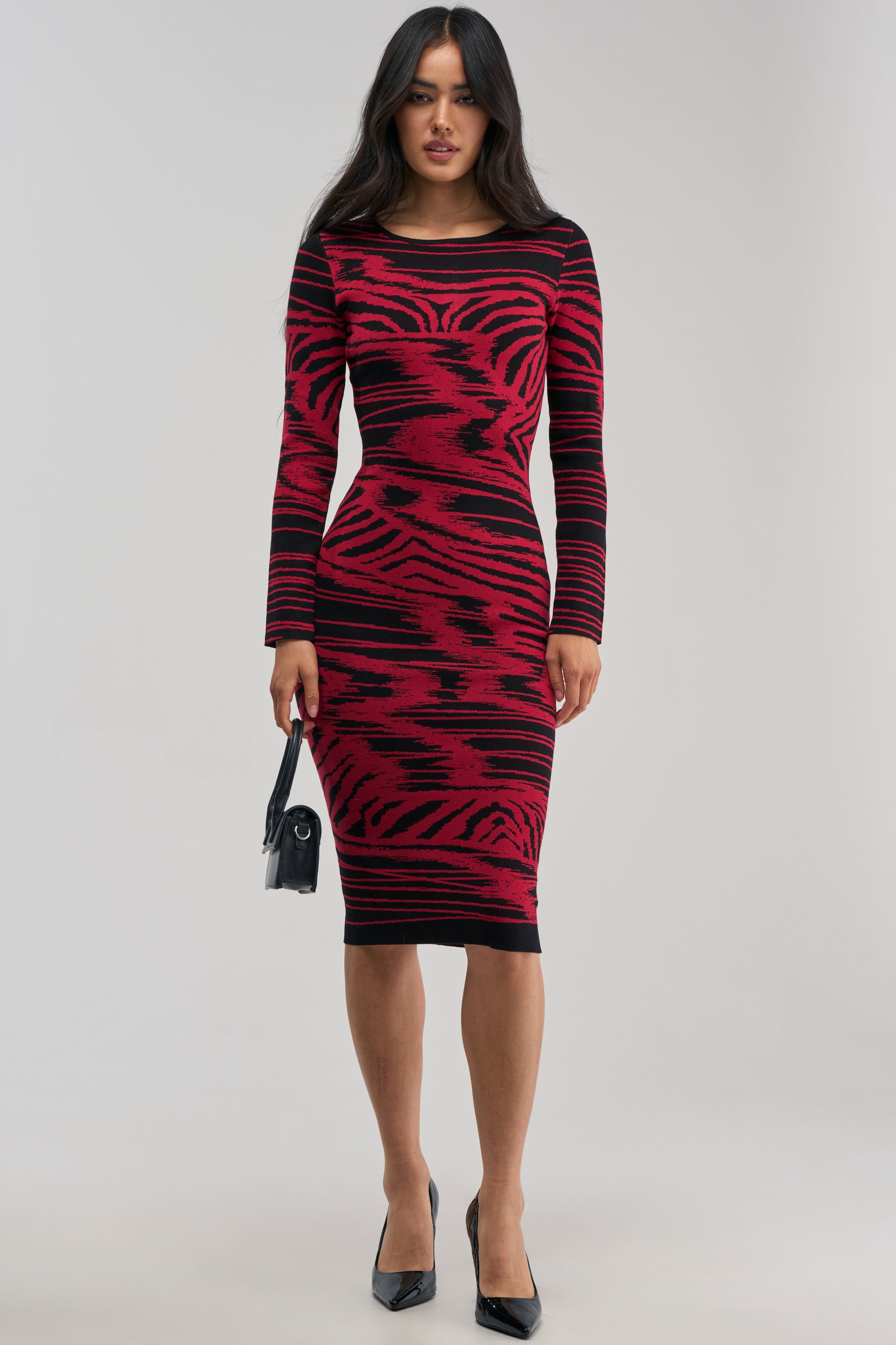 Wearing the HEART EYES ONLY KNIT MIDI DRESS—a fitted, long-sleeve bodycon midi dress featuring an eye-catching red and black abstract print—a woman stands against a plain light background. She completes this striking winter date night look with a small black handbag and black pointed-toe heels.