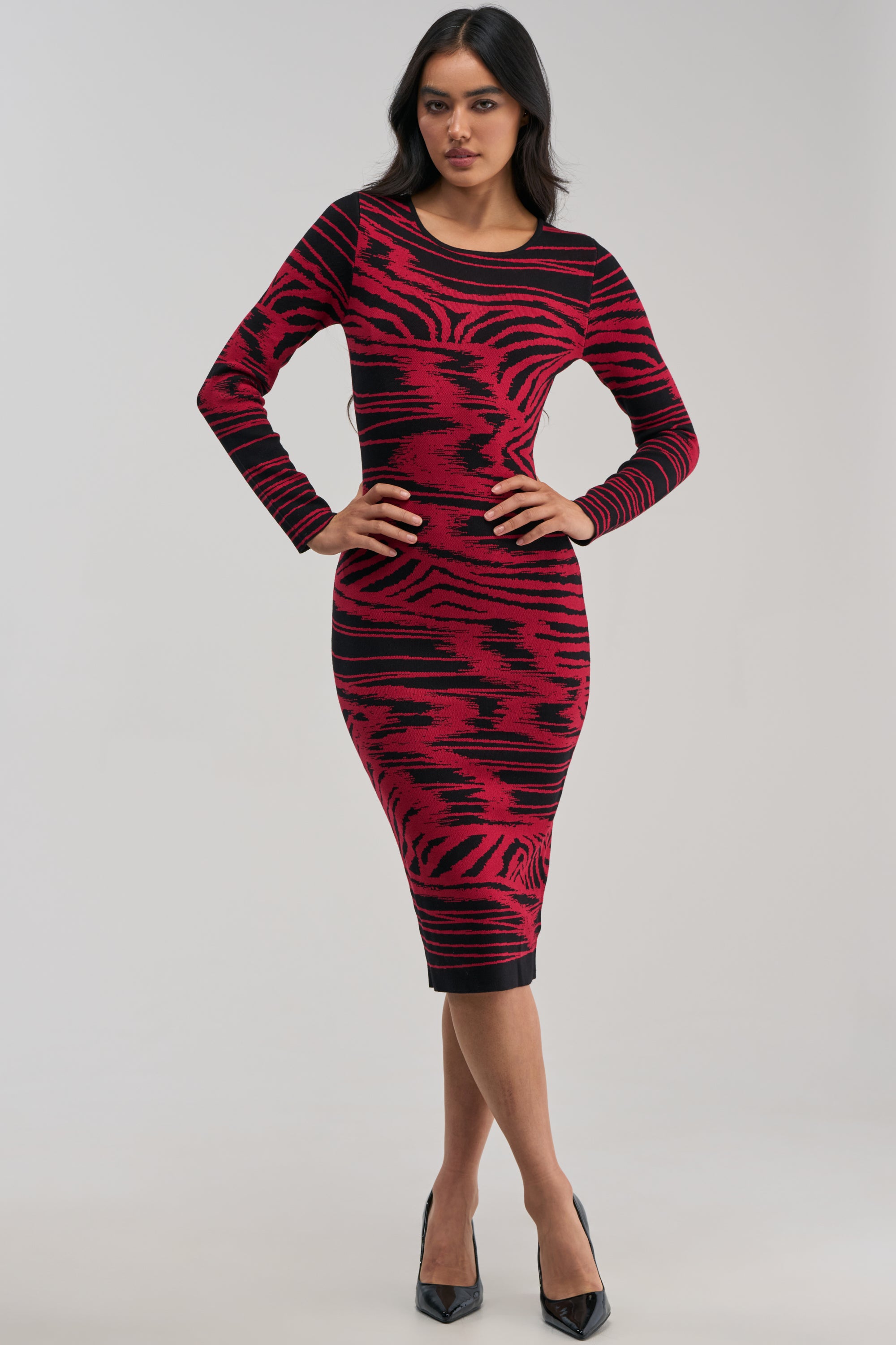 Wearing the HEART EYES ONLY KNIT MIDI DRESS—a fitted, long-sleeve red and black bodycon midi dress featuring an abstract knit pattern—she stands confidently with hands on her hips against a plain light background, paired with black high heels. The perfect winter date night look.