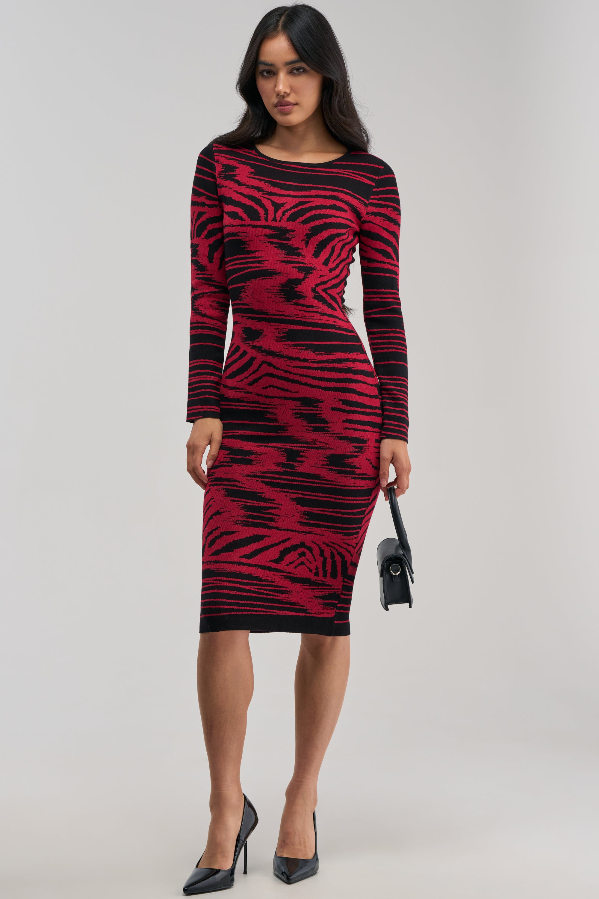 A woman models the HEART EYES ONLY KNIT MIDI DRESS—a fitted, long-sleeve bodycon midi dress featuring a striking red and black tiger stripe pattern. She pairs the look with a small black handbag and black high heels, standing against a plain light background.