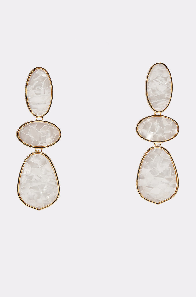 The TRIPLE DROP EARRINGS IN WHITE feature three irregular, white, semi-translucent stones set in gold-tone teardrop drops, vertically stacked against a plain white background.