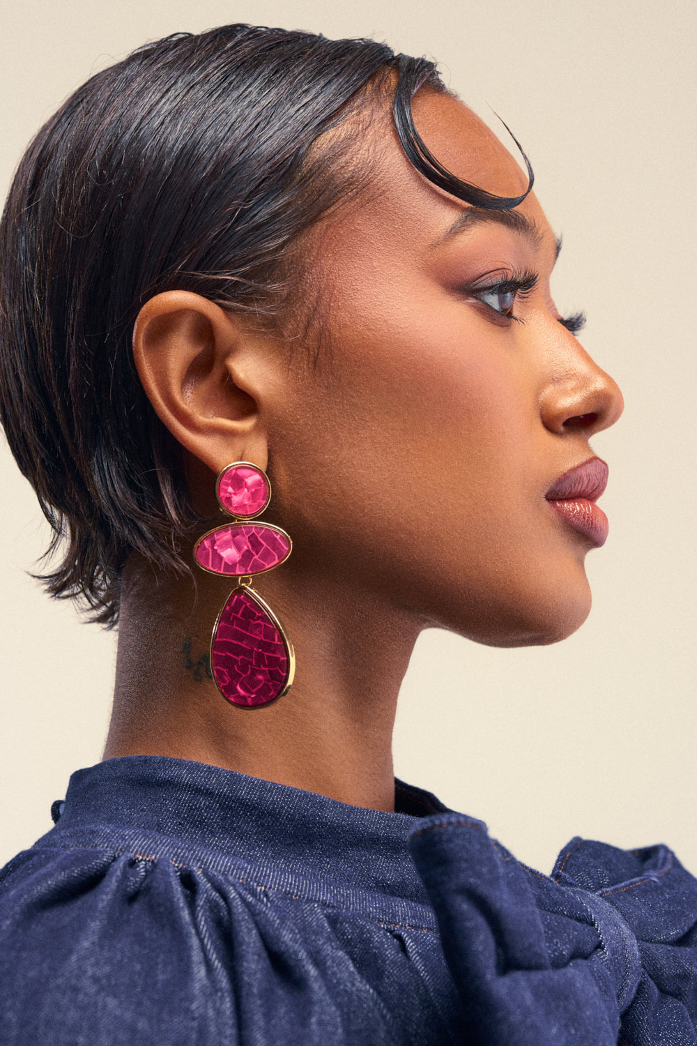 A woman in profile showcases the TRIPLE DROP EARRINGS IN PINK—striking pink teardrop earrings ideal for summer parties—paired with a navy blue top and subtle makeup. Her hair is short and sleek, revealing a small tattoo just behind her ear.