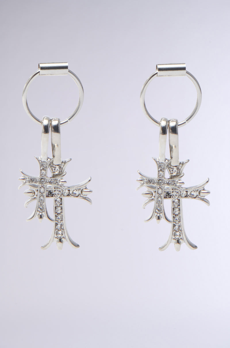 The CROSS CHARM EARRING features two silver metallic hoops with cross charms encrusted with rhinestone crystals, set against a plain gray background. 0