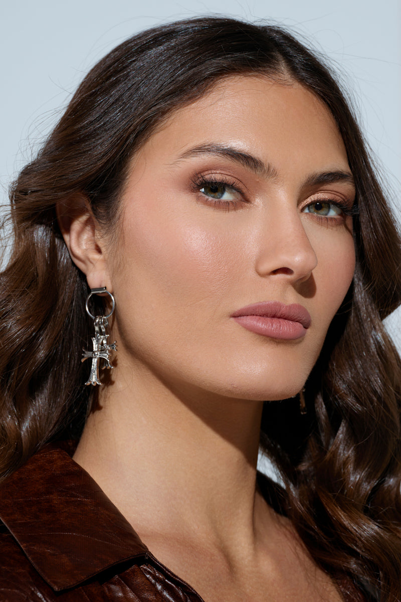 A woman with long, dark wavy hair wears a brown top and CROSS CHARM EARRING while looking at the camera. The plain background emphasizes her natural makeup and confident expression, accentuating the rhinestone-embellished details that add sparkle. 1