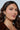 A woman with long, dark wavy hair wears a brown top and CROSS CHARM EARRING while looking at the camera. The plain background emphasizes her natural makeup and confident expression, accentuating the rhinestone-embellished details that add sparkle.