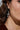 A person wears silver metallic CROSS CHARM EARRINGS, featuring intricate branches and rhinestone embellishments. These earrings hang from a hoop, beautifully complementing the person's long brown hair while the image focuses on their dazzling charm against the side of their face.