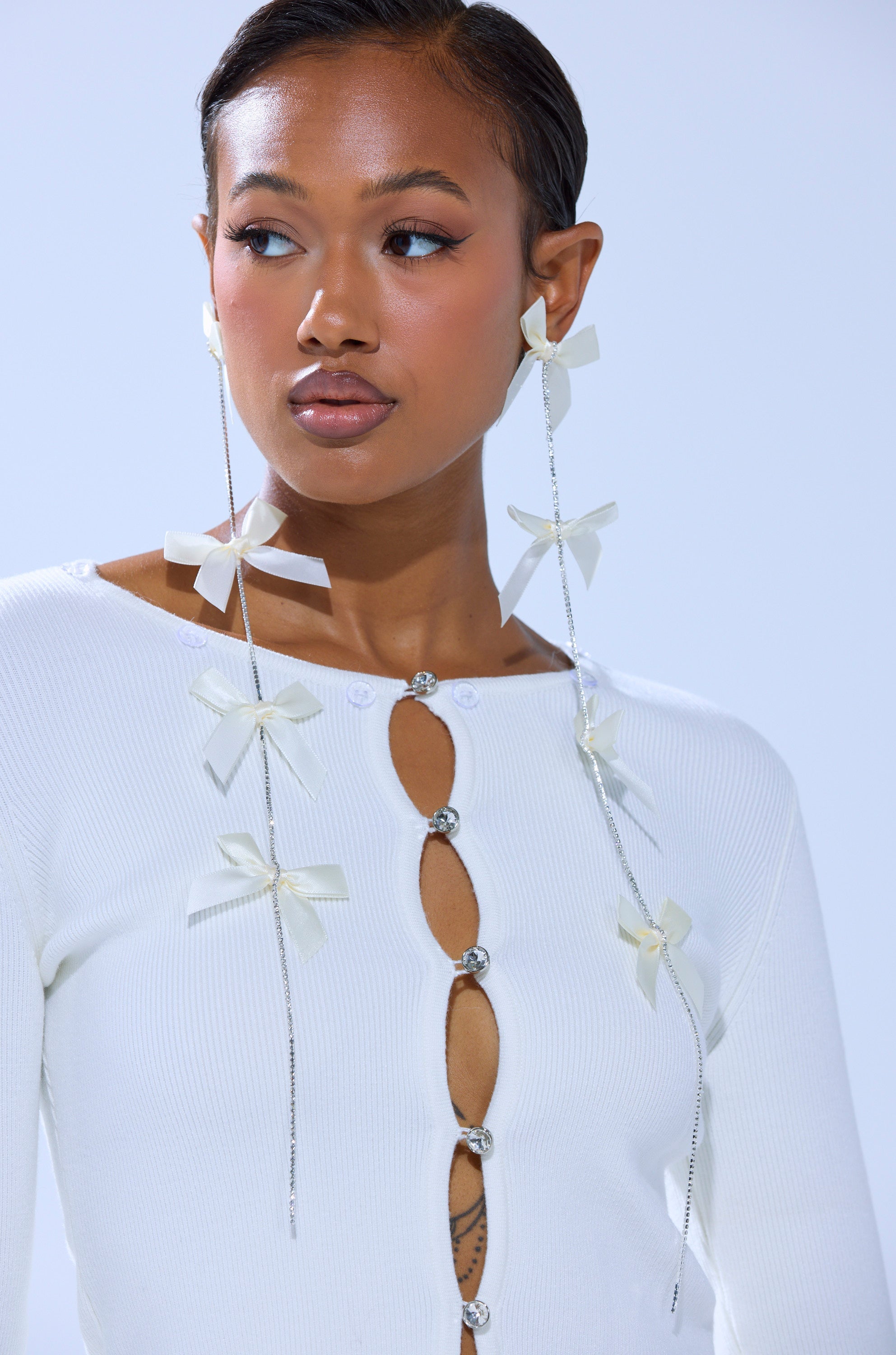 A woman with short slicked-back hair and subtle makeup wears a white top with jeweled buttons and bows, paired with COQUETTE DREAMING EARRINGS—long rhinestone bow earrings. The background is light blue. 2