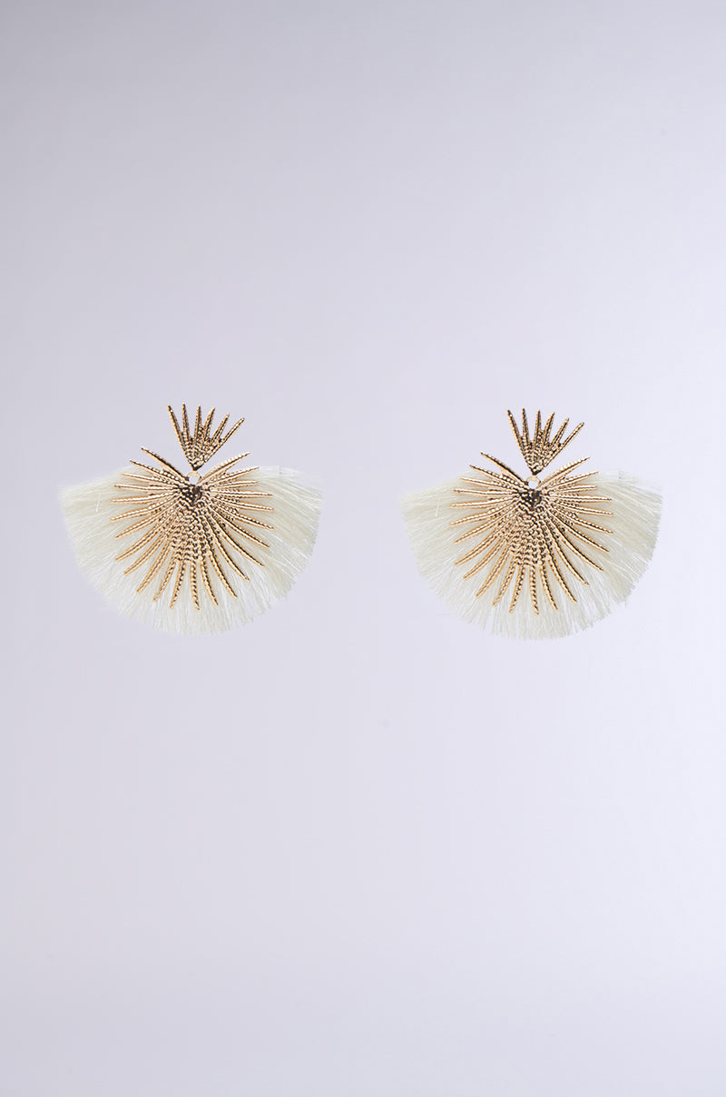 The TULUM EARRING is a pair of resort-glam statement earrings with gold sunburst designs and white fan-shaped fringes, shown on a light gray background. 0