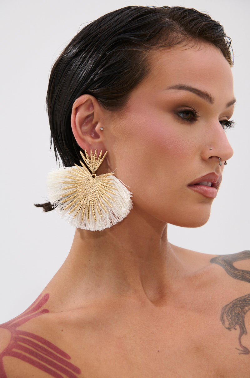 A person with short, wet-look dark hair models the TULUM EARRING—a bold gold and white fan-shaped piece—evoking resort glamour. Multiple ear piercings, a nose ring, natural makeup, and a shoulder tattoo complete the stylish look. 1