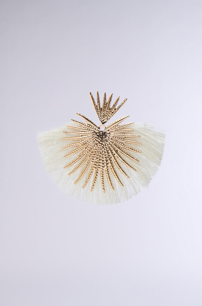 The TULUM EARRING features a gold-toned sunburst design with textured rays and white fringe, creating a resort-glam statement against a light gray background. 3