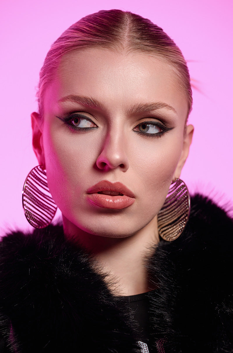 A woman with sleek, pulled-back hair and bold makeup gazes to the side. She wears the MAIN MOMENT BLING HOOP EARRING, adorned with rhinestones, along with a black faux fur collar. The background is a soft pink, casting a gentle glow.