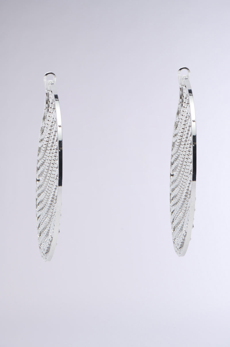 A close-up shot captures the MAIN MOMENT BLING HOOP EARRINGS against a plain gray background. These earrings showcase an intricately detailed woven pattern on their metallic base, appearing suspended in mid-air and shimmering with elegant craftsmanship.