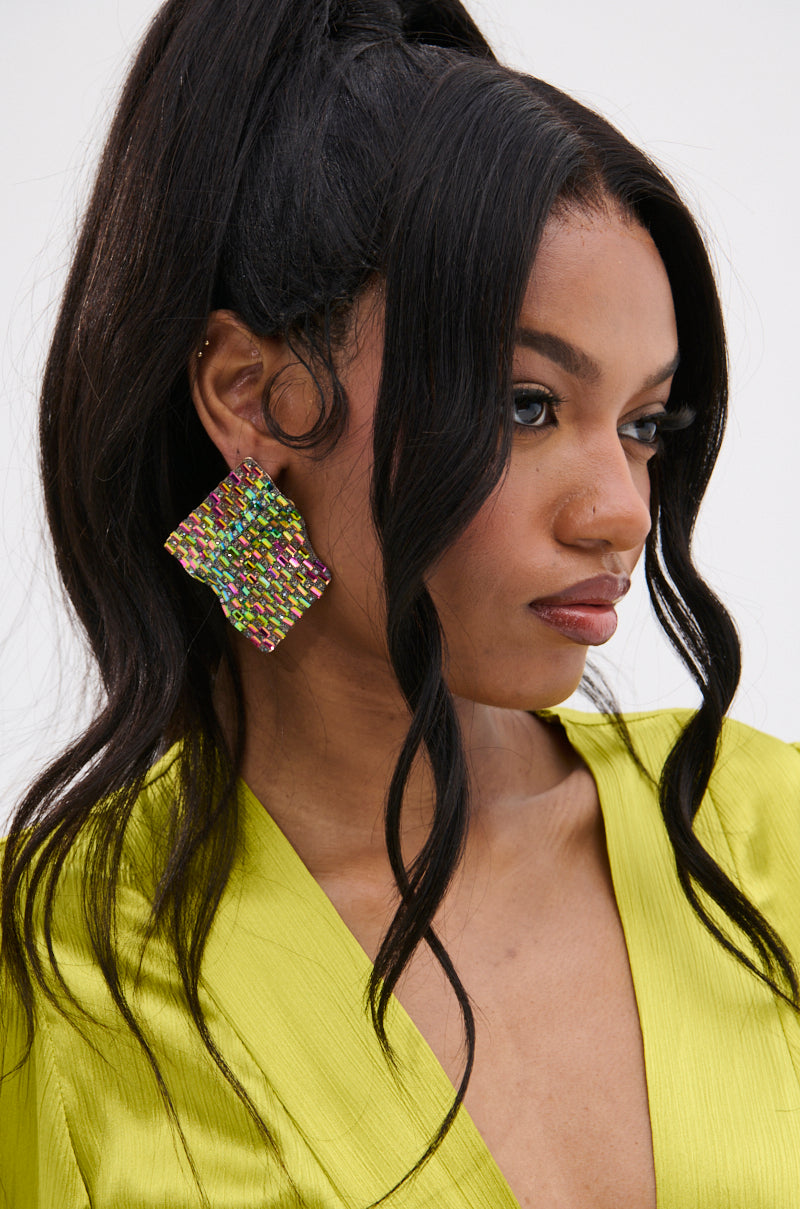 A woman with long, wavy black hair in a high ponytail wears a lime green top and the SHINE BRIGHT EARRING—oversized and adorned with multicolor rhinestones. She has a nose piercing and glances slightly to the side. 1