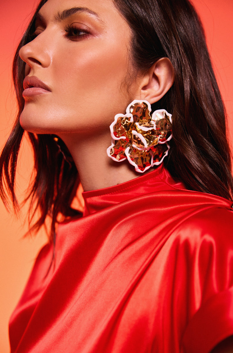 A woman in a red satin top poses against an orange gradient background, wearing the IN BLOOM EARRING—large gold floral statement earrings with a hammered texture. She has shoulder-length dark hair and natural makeup.