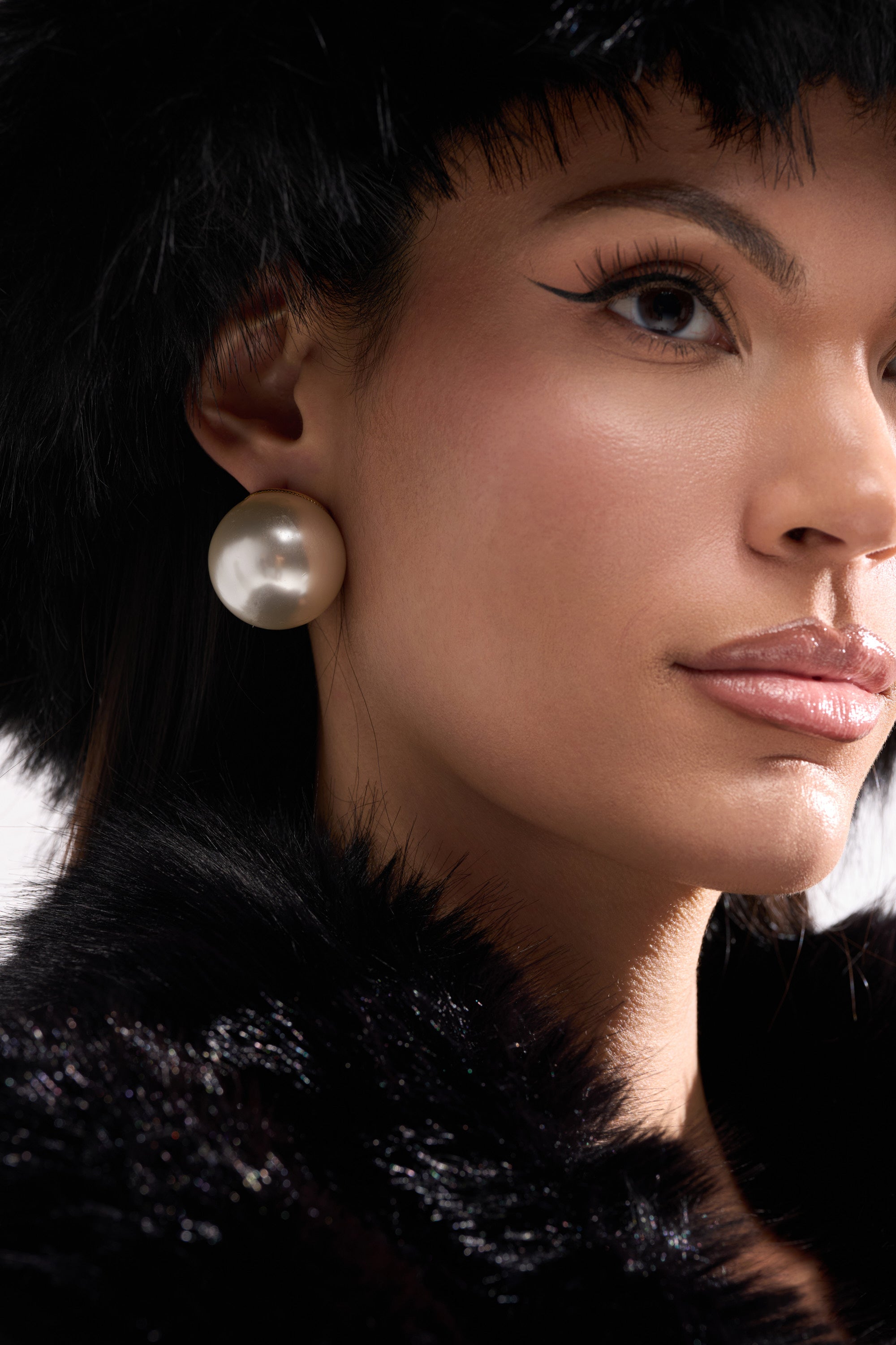 A close-up of a woman wearing the TIFF EARRING, a pair of statement pearl earrings, along with a black fur hat and coat. She has winged eyeliner and glossy lips, gazing confidently to the side. 3
