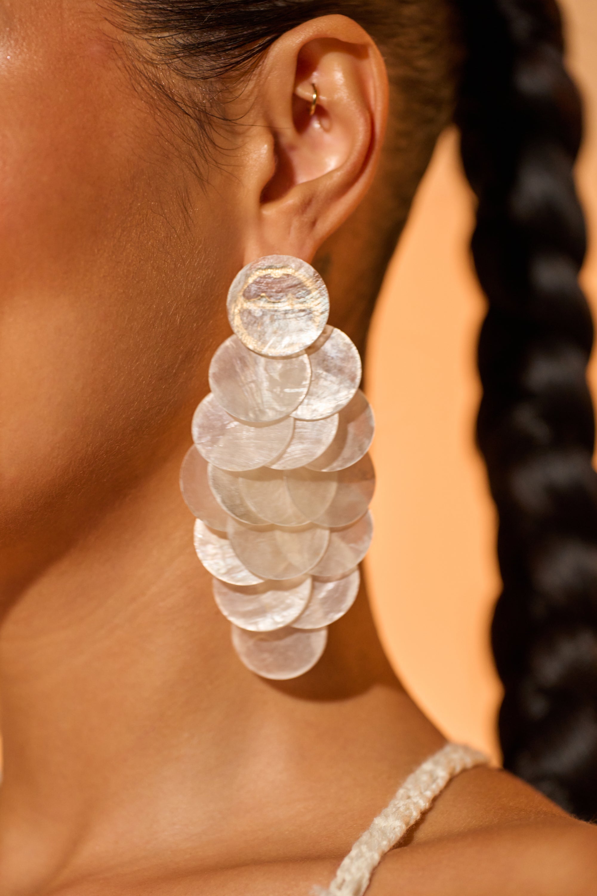 Close-up of a person wearing the GIMME MY SHELLS EARRING, featuring overlapping, iridescent translucent discs. The person’s hair is styled in braids and they also wear a gold ear cuff on the upper ear. 0