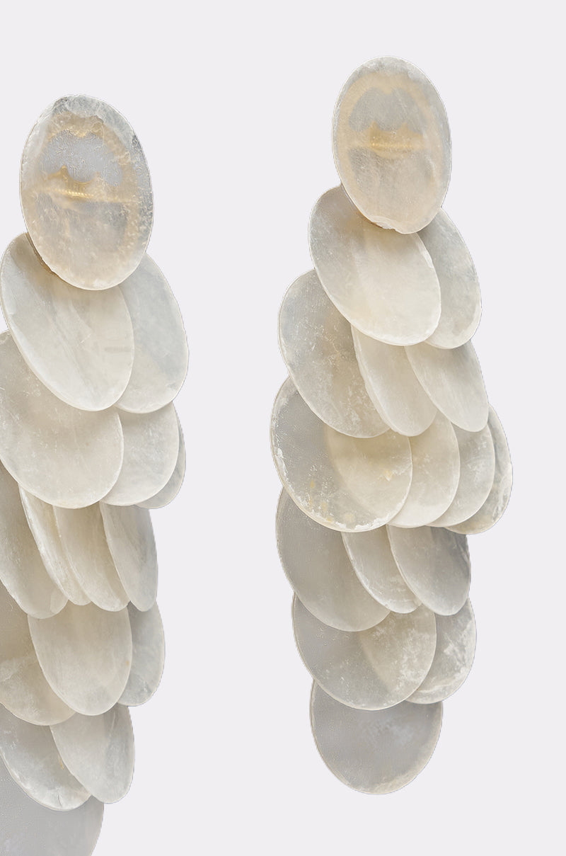 The GIMME MY SHELLS EARRING features layered, overlapping translucent white discs with a subtle mother-of-pearl finish, cascading elegantly against a plain light background. 3