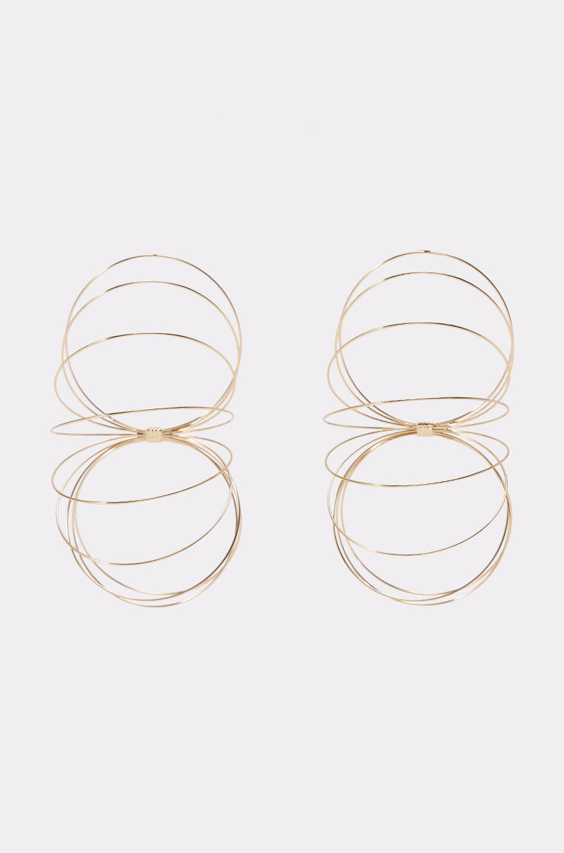 The GOLD SLINKY EARRINGS are a pair of large, gold wire hoop statement earrings featuring interlocking circular designs, displayed on a plain white background.