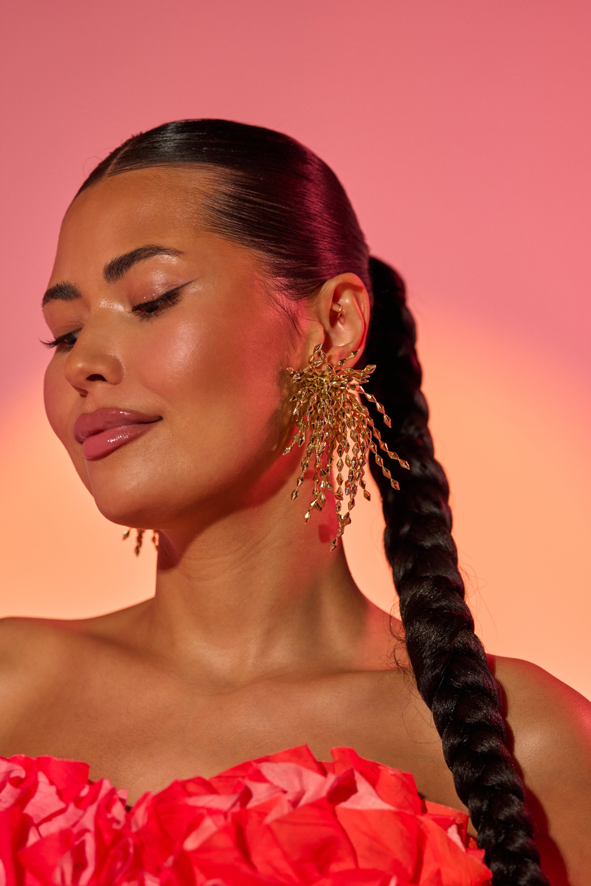 A woman with long, dark braided hair wears the striking THATS DRAMA EARRING and a bright textured red top, posing with her head tilted and eyes closed against a pink and orange gradient background.