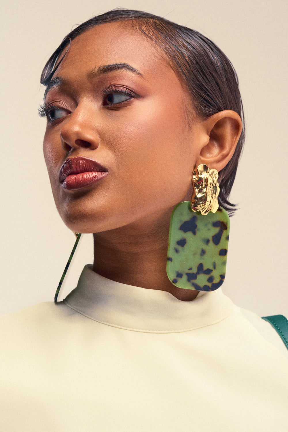 A woman with short, sleek hair wears a cream top and the bold CAMO CRUSH RESIN EARRINGS—one gold, one green with black spots—embodying striking summer style. She gazes confidently to the side against a neutral background, her look elevated by subtle gold accents.