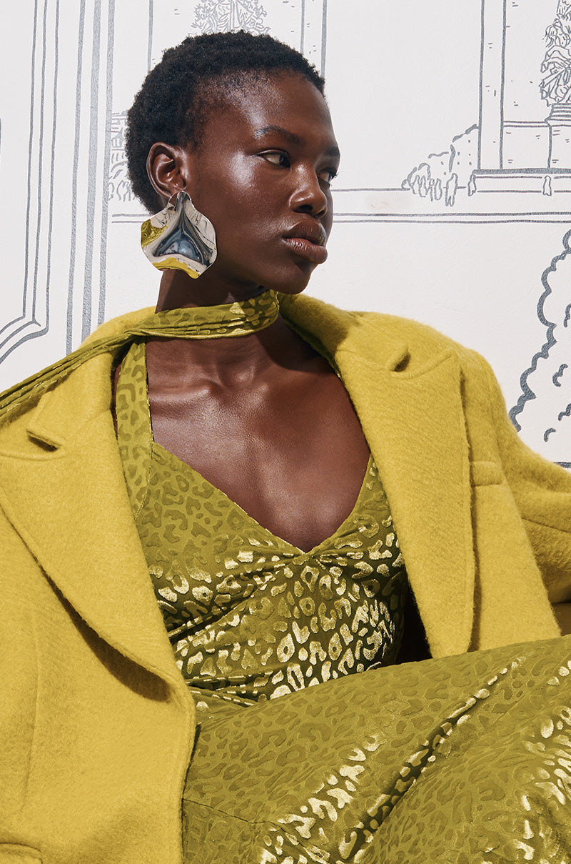 A person wearing a stylish green patterned dress and oversized coat poses against a monochrome illustrated backdrop. They have short hair and are accessorized with the large, abstract TAKE ME TO PARIS EARRINGS, adding an avant-garde touch to their ensemble.
