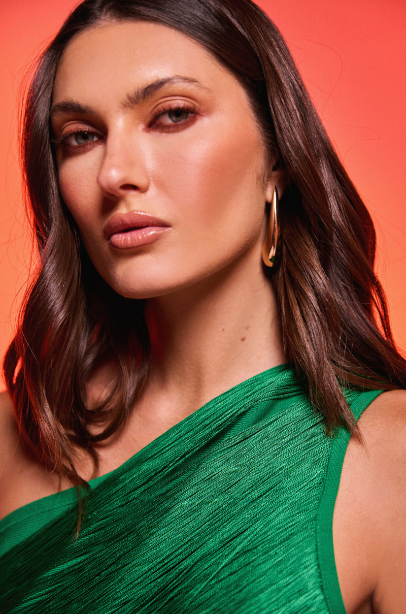A woman with wavy brown hair, wearing a green textured one-shoulder top and the IN THE WIND EARRING in gold-tone, poses against a warm red-orange background.