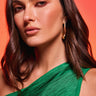 A woman with wavy brown hair, wearing a green textured one-shoulder top and the IN THE WIND EARRING in gold-tone, poses against a warm red-orange background.