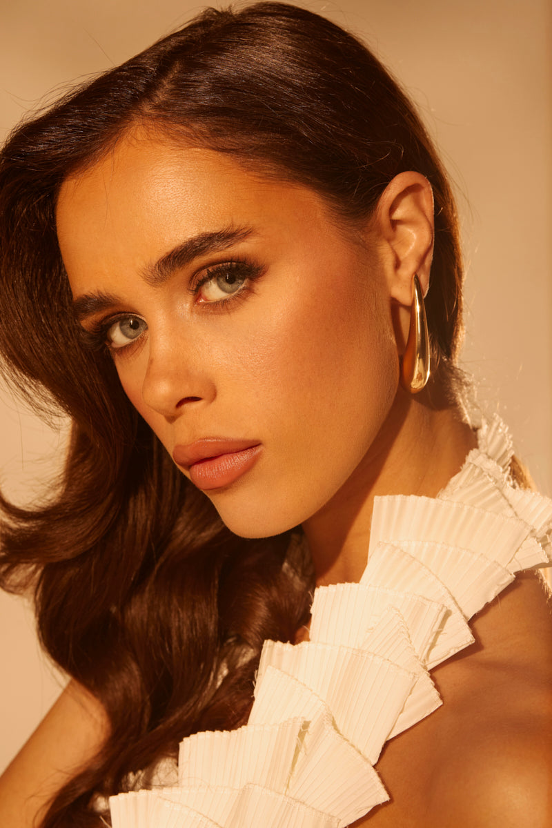 A woman with long brown hair and green eyes looks at the camera, her facial features highlighted by warm, soft lighting. She wears a white ruffled top and gold-tone IN THE WIND EARRING featuring an abstract silhouette design.