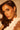 A woman with long brown hair and green eyes looks at the camera, her facial features highlighted by warm, soft lighting. She wears a white ruffled top and gold-tone IN THE WIND EARRING featuring an abstract silhouette design.
