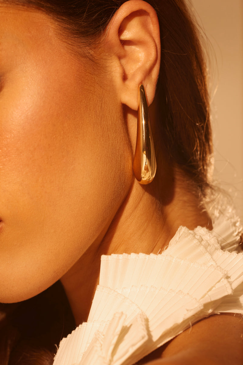 Close-up of a woman's ear wearing the IN THE WIND EARRING—a large, gold-tone teardrop design. She has warm-toned skin, a white pleated top, and soft golden lighting highlights the earring's abstract silhouette.