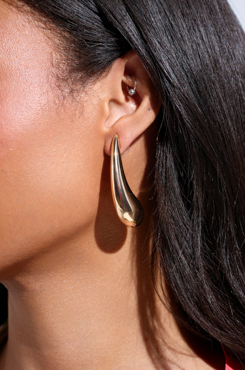 Close-up of a woman's ear wearing the IN THE WIND EARRING, a large abstract gold silhouette, paired with a small silver hoop. Her dark hair is tucked behind her ear and her skin glows in natural light. 4