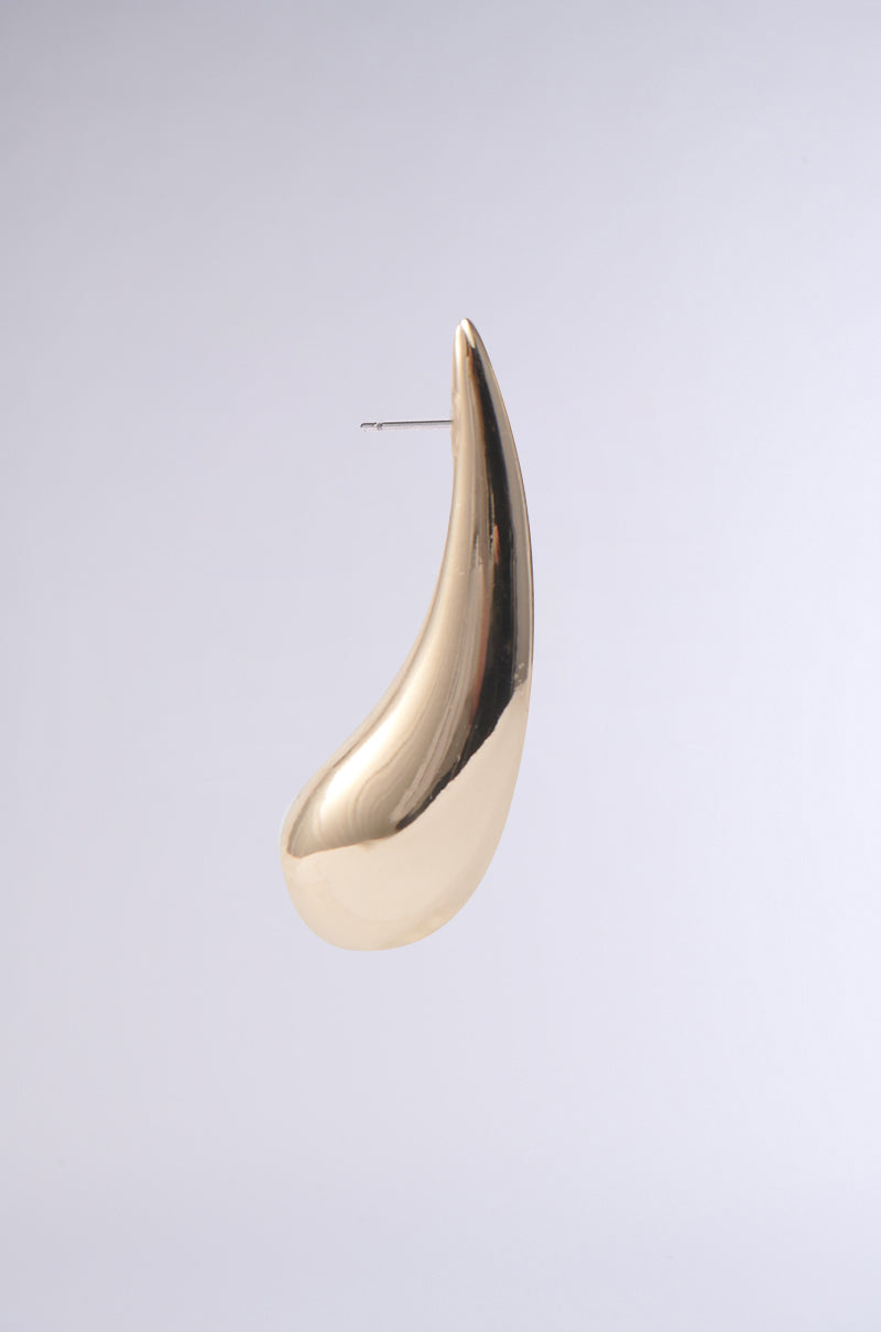The IN THE WIND EARRING, featuring a smooth gold-tone finish and an elongated abstract teardrop silhouette, is displayed against a plain light gray background.