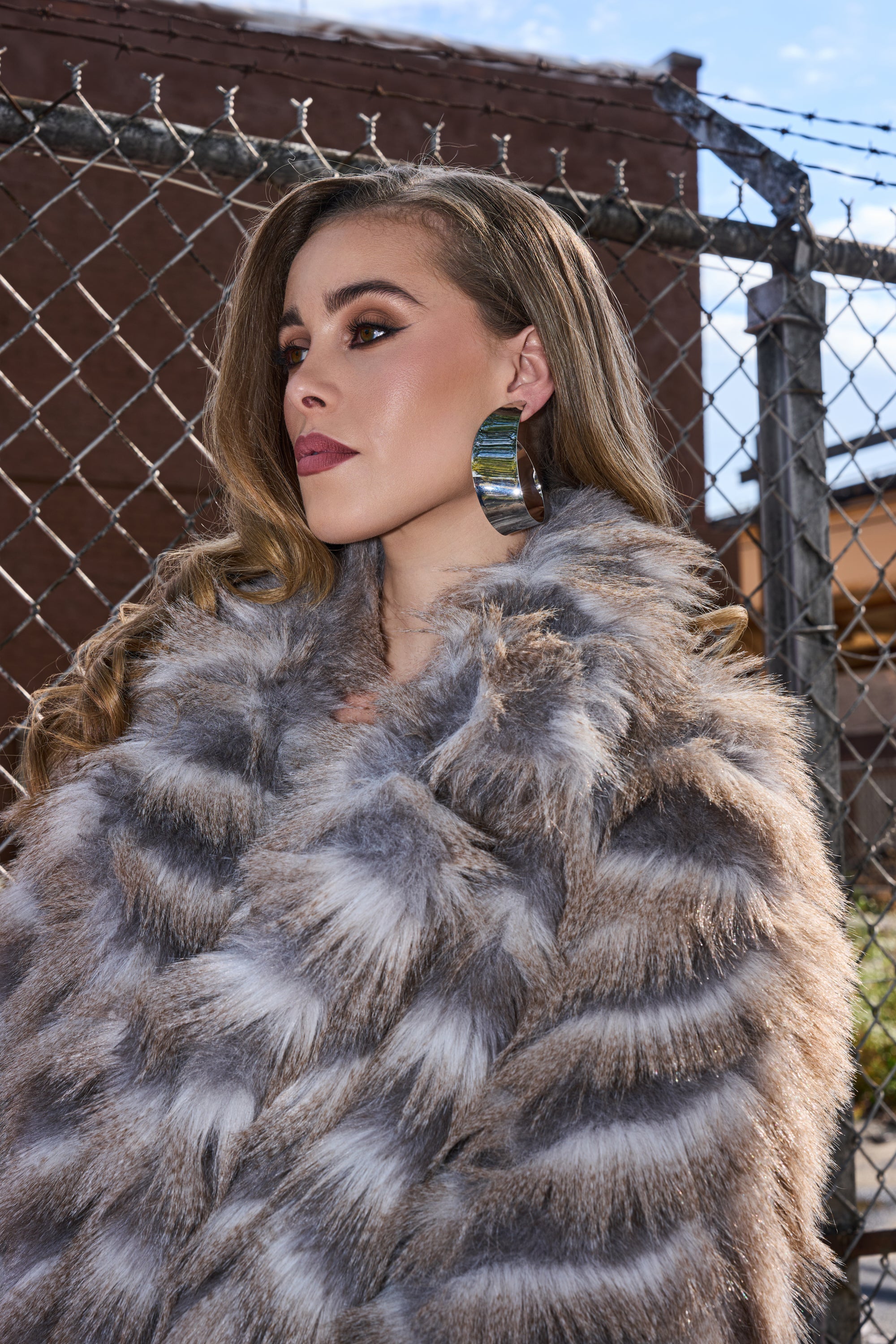 Wearing bold makeup and metallic silver HOOPIN EARRING, a woman with long wavy hair and a luxurious gray fur coat stands by a chain-link fence and brick wall on a sunny day. 5