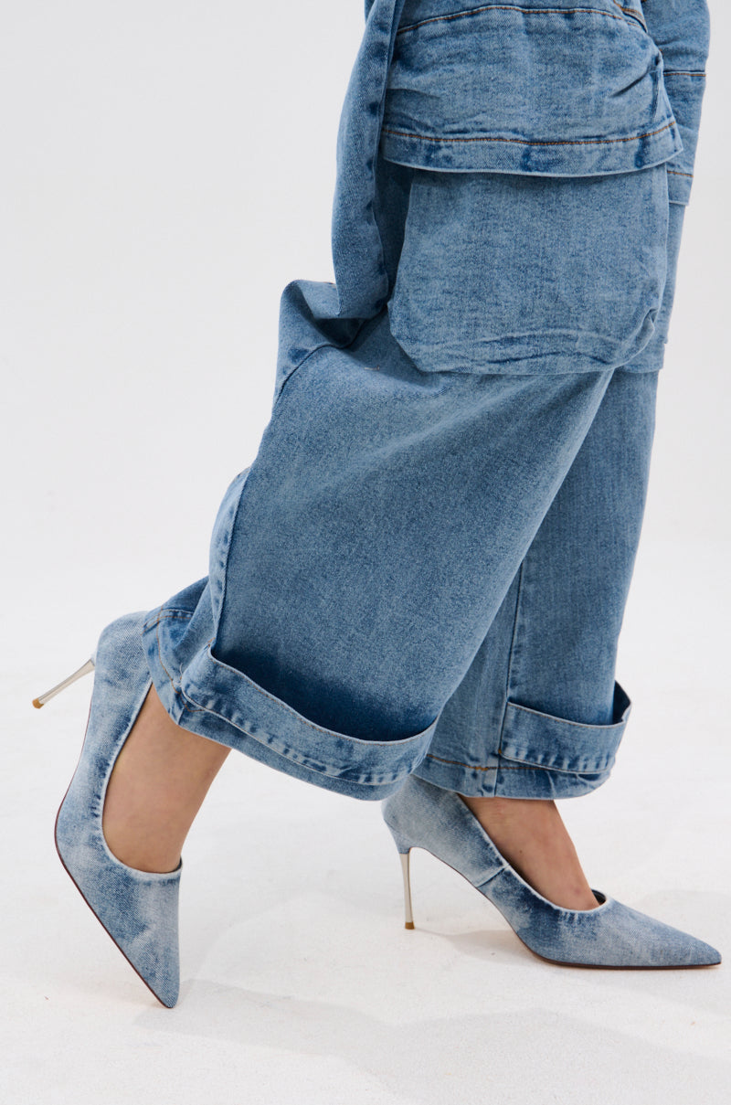 Against a plain light background, a person wears loose-fitting, cuffed blue jeans and the AZALEA WANG KEATON DENIM STILETTO PUMP with pointed toes and high heels.