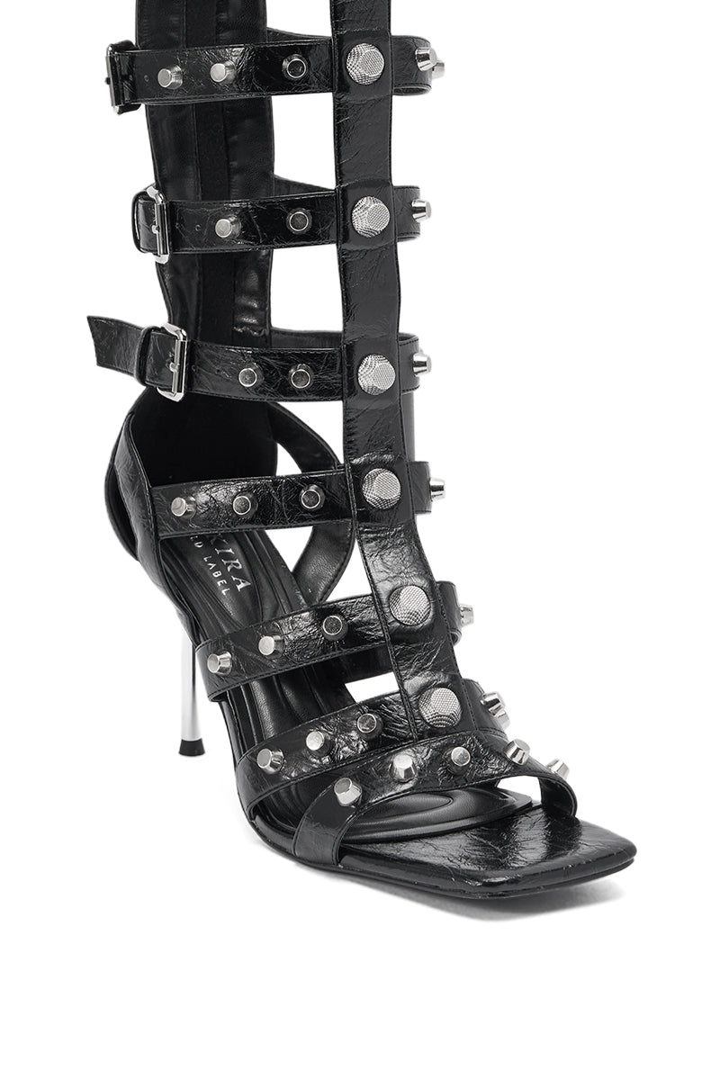 The AKIRA GOLD HARDWARE EMBELLISHED BLACK GLADIATOR SANDAL features multiple leather straps, gold hardware studs, a square toe, and a striking metallic stiletto heel for a bold look. 6