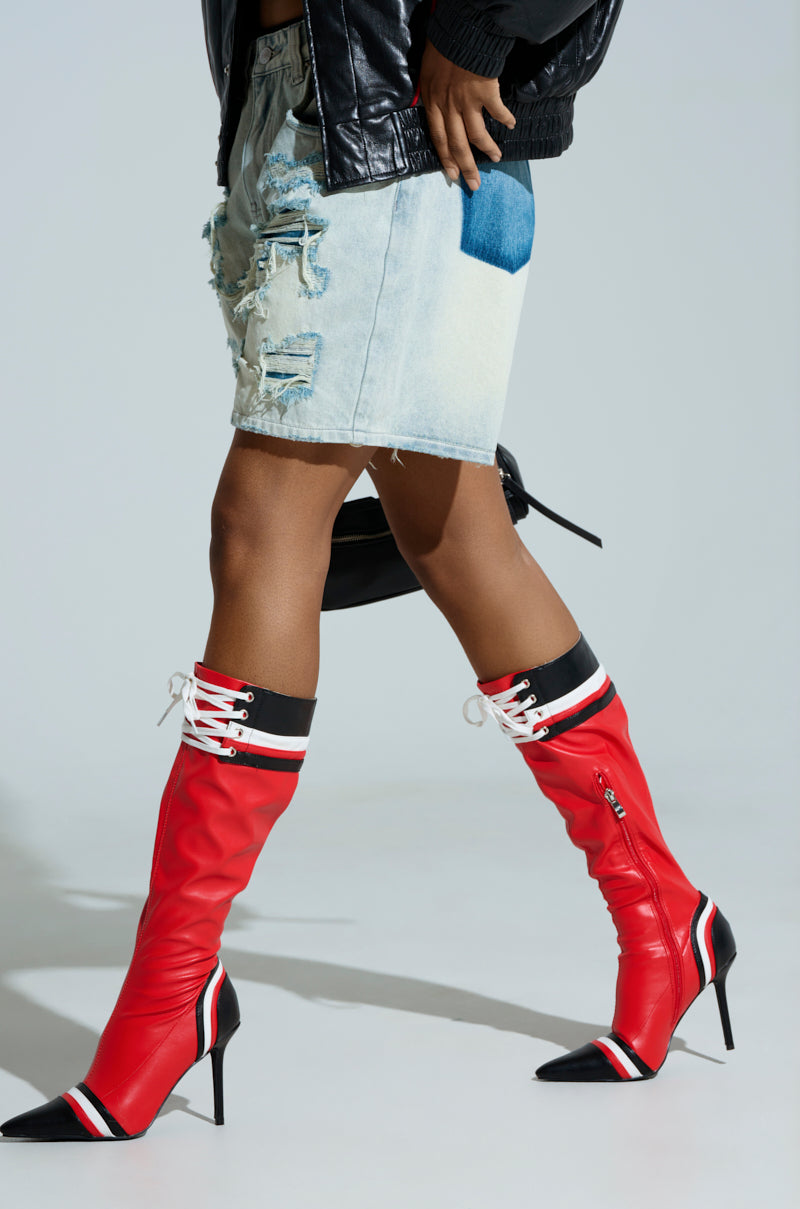 Wearing AZALEA WANG KELCIE red stiletto boots with white laces and black tips, a distressed denim skirt, and a sleek black faux leather jacket, the person clutches a small black handbag against a plain gray backdrop.