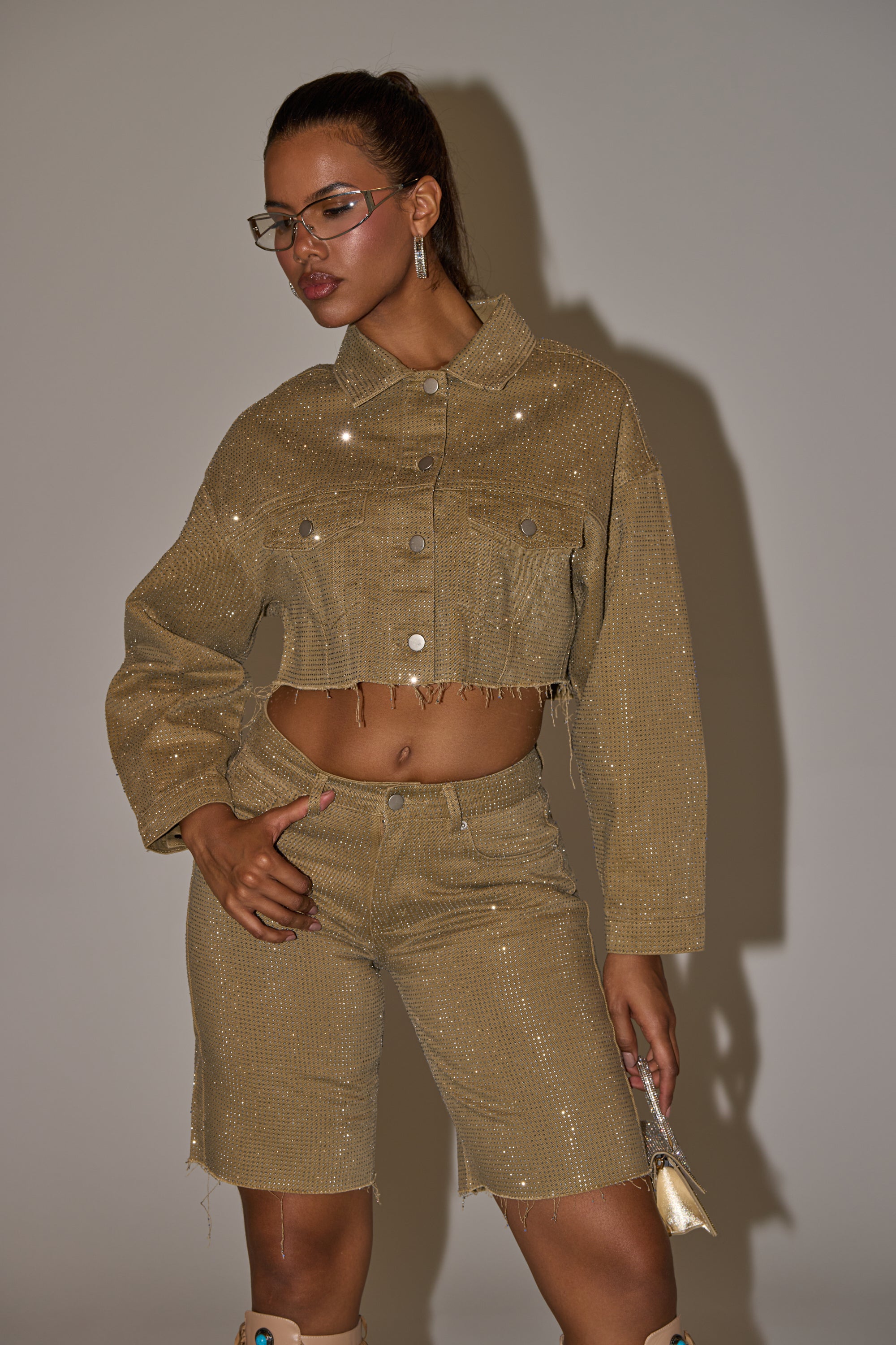 A woman poses against a plain background wearing the CHECK OUT THE BLING DENIM JACKET with rhinestone embellishments and matching knee-length shorts. She accessorizes with glasses, earrings, and a small metallic clutch in her left hand. 2