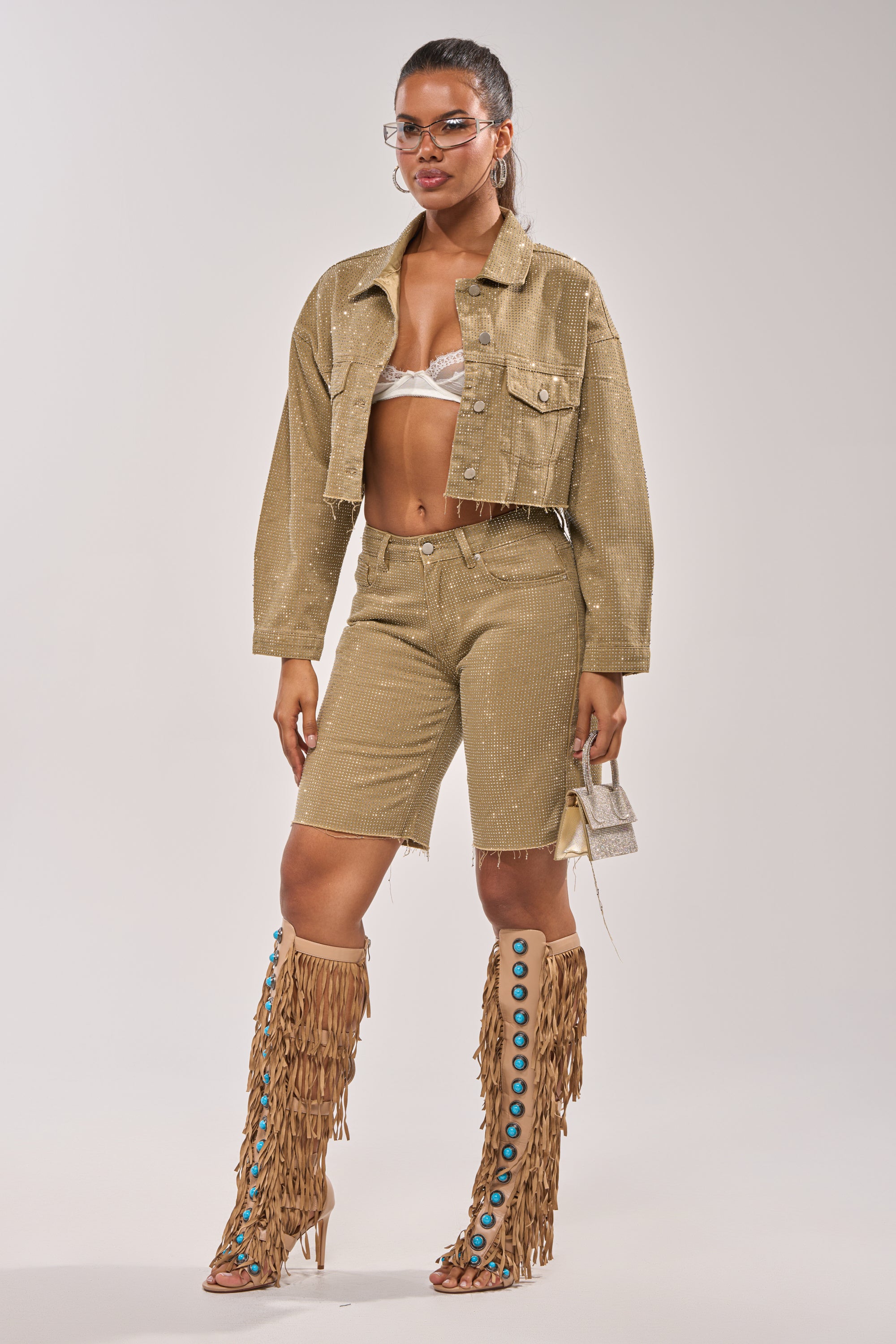 A woman poses against a light background wearing the CHECK OUT THE BLING DENIM JACKET, matching high-waisted shorts, a white bralette, tall tan fringe boots with blue accents, and holds a small silver handbag. 11