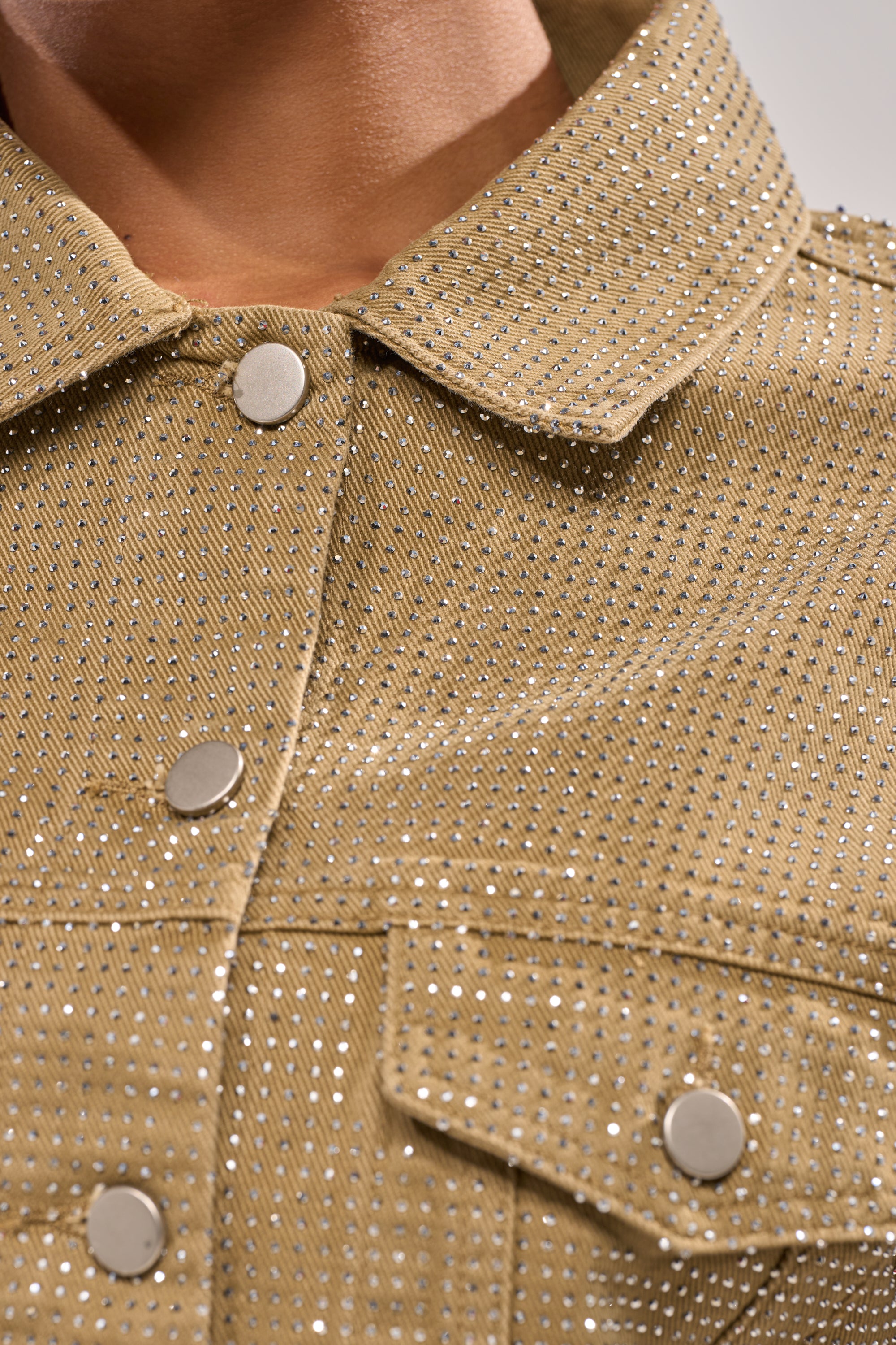Close-up of the CHECK OUT THE BLING DENIM JACKET—a khaki piece featuring rhinestones, metallic studs, silver snap buttons, and a textured pattern that sparkles in the light. Worn by a person with collar visible. 12