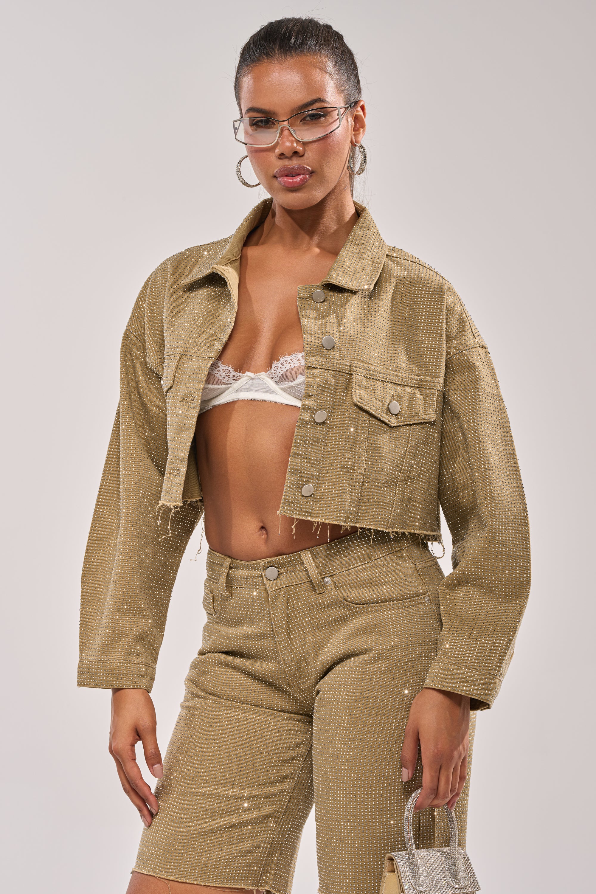 A woman wearing the CHECK OUT THE BLING DENIM JACKET with matching shorts, a white lace bralette, clear eyeglasses, and a small silver handbag stands against a plain light background. 3