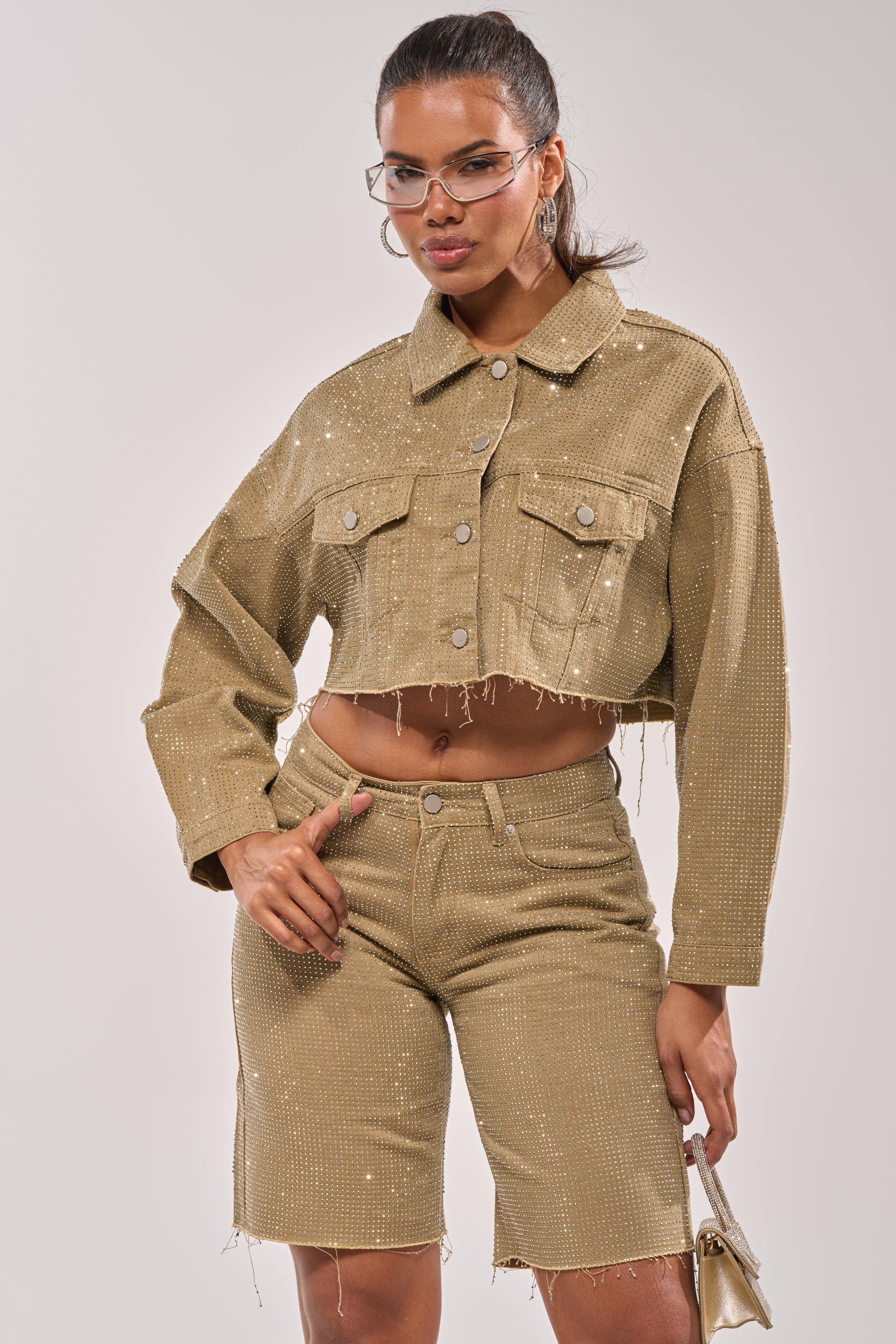 A woman wearing the CHECK OUT THE BLING DENIM JACKET and matching embellished shorts poses confidently. She has glasses, her hair in a ponytail, and carries a small metallic purse. 5