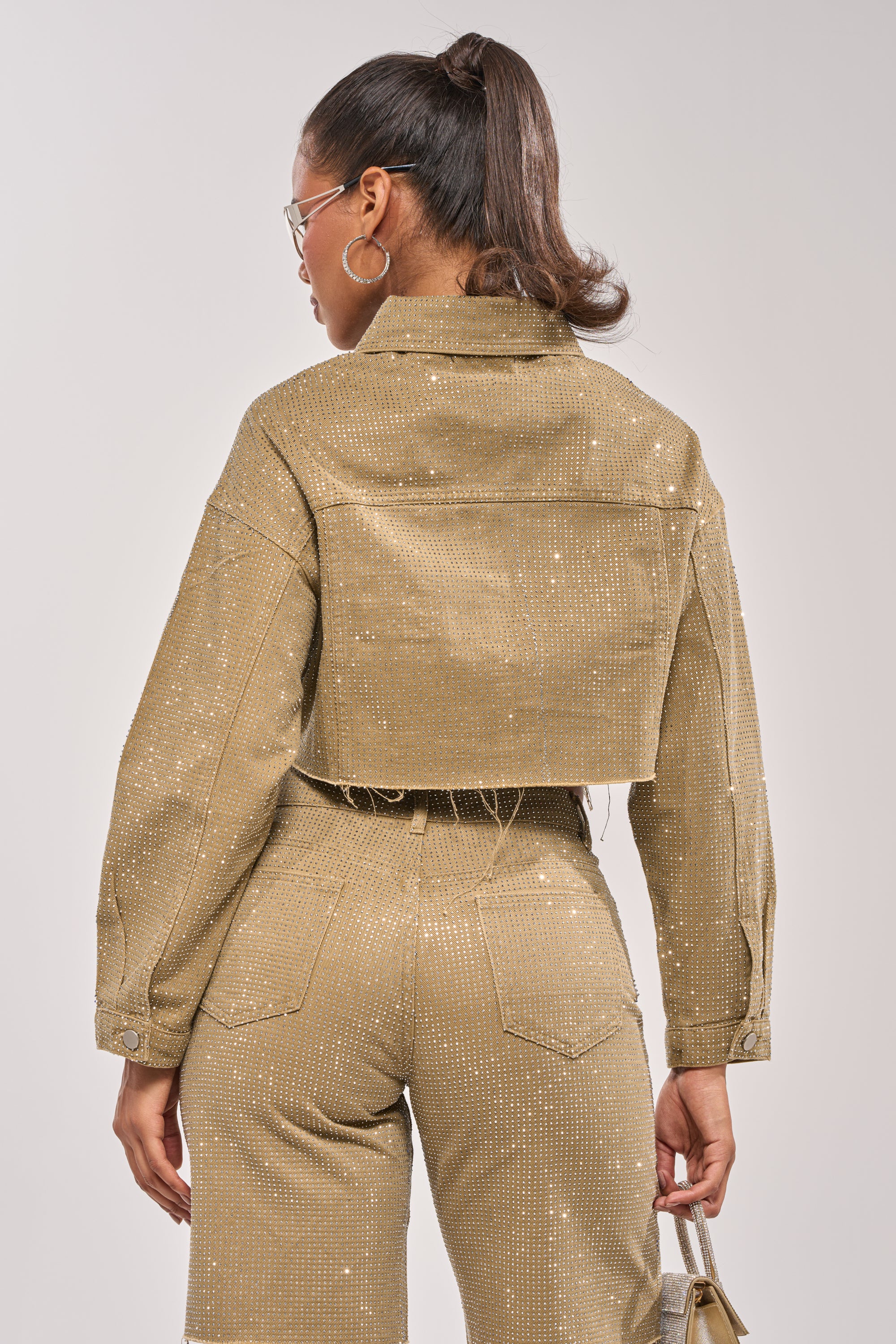 Woman with a high ponytail wears the CHECK OUT THE BLING DENIM JACKET and matching high-waisted pants, holding a small gold purse, seen from the back against a plain light background. 8