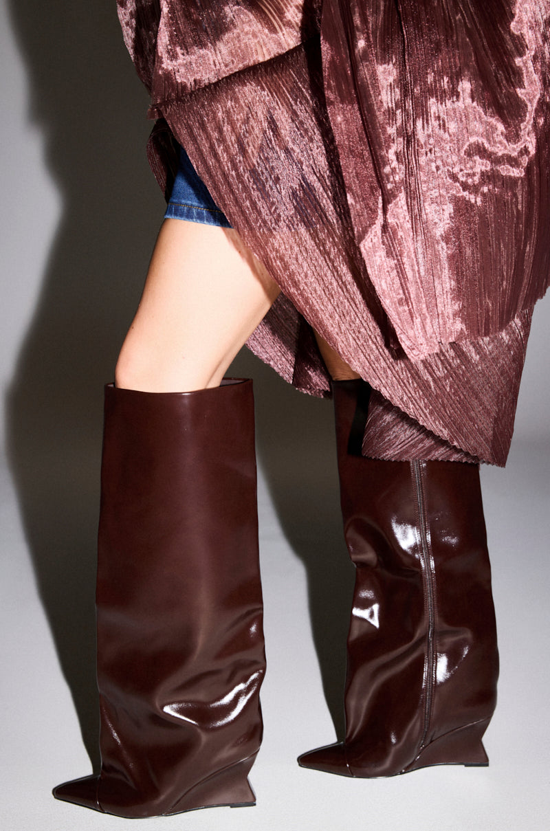 A person wears AZALEA WANG KENYA BROWN KNEE HIGH WEDGE BOOTS with pointed toes and a brown pleated skirt, standing on a light gray floor. Only the lower half of their body is visible.