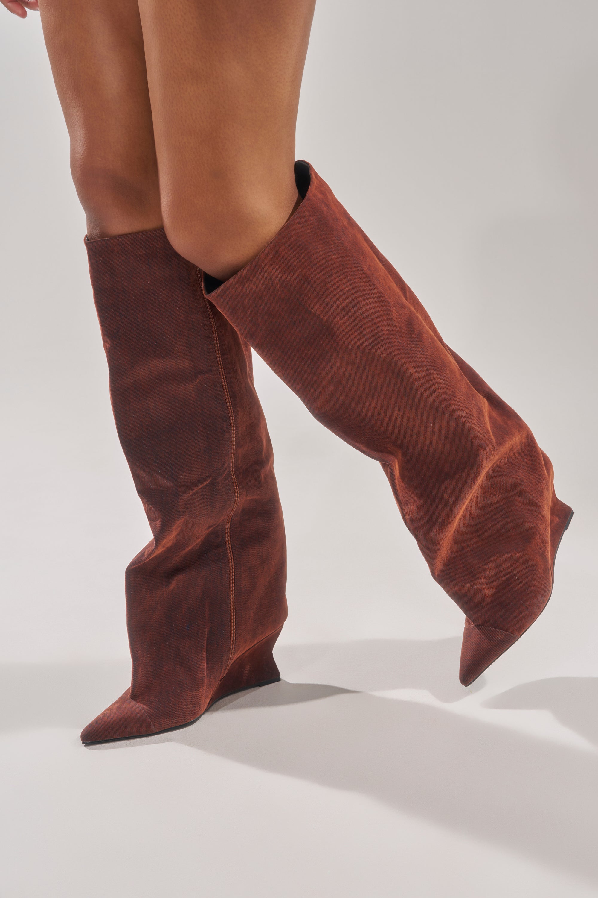 A person models the AZALEA WANG KENYA BURNT ORANGE VELVET KNEE HIGH WEDGE BOOT, standing with one leg slightly bent to highlight the rich velvet texture and striking shape against a plain background.