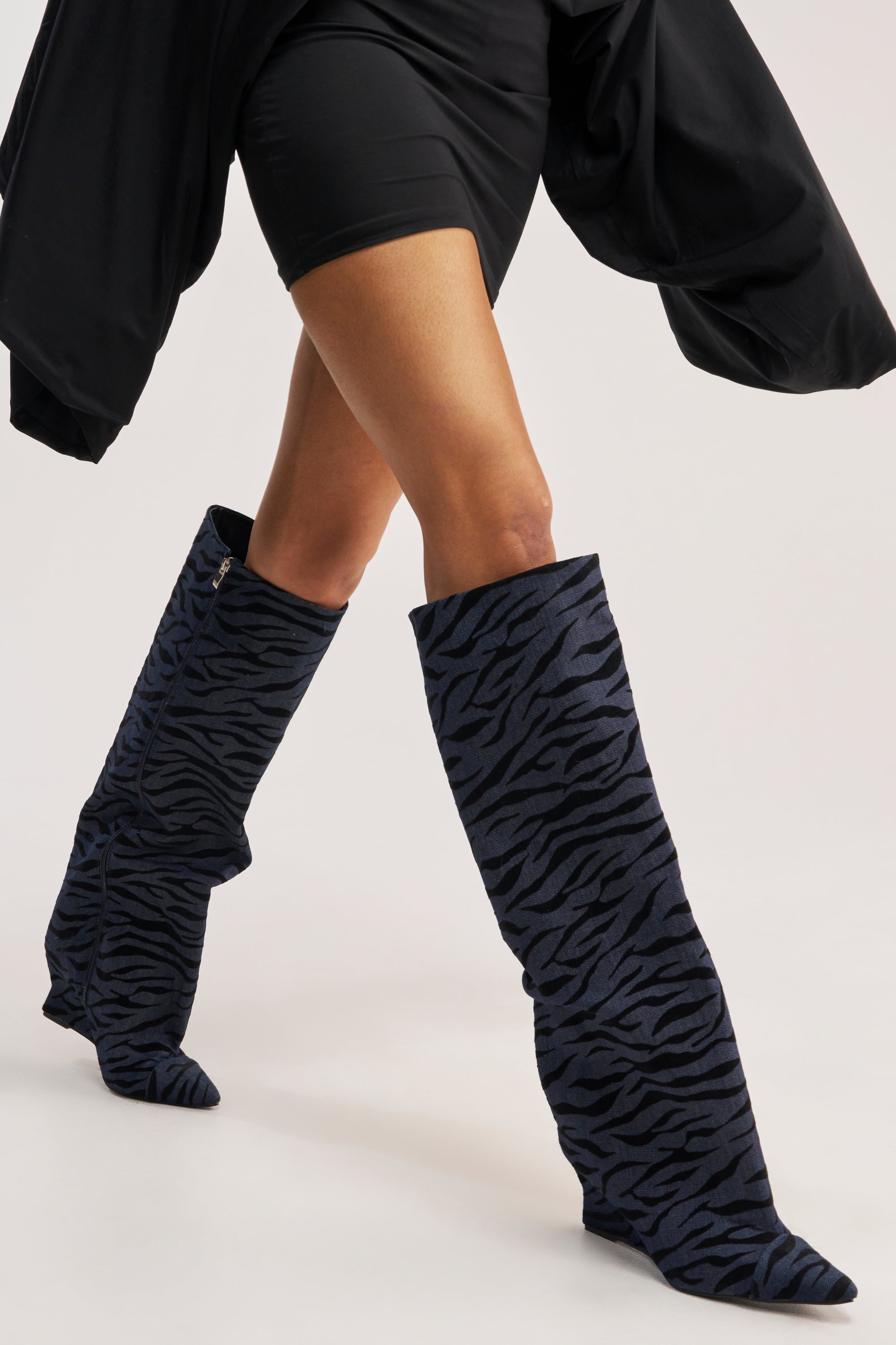 A person wearing a black dress and the AZALEA WANG KENYA DENIM TIGER WEDGE BOOT walks forward, emphasizing their legs against a plain light background.